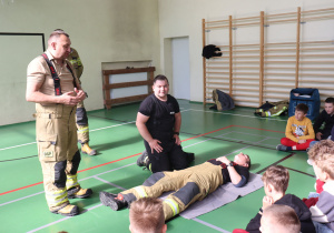 Na sali gimnastycznej odbywa się pokaz udzielania pierwszej pomocy. Dwóch strażaków, jeden stojący, drugi klęczący, prowadzą instruktaż dla grupy dzieci siedzących na podłodze. Trzeci strażak leży na macie, symulując osobę poszkodowaną. W tle widoczne są drabinki gimnastyczne, sprzęt ratowniczy i kurtki. Dzieci uważnie obserwują demonstrację.