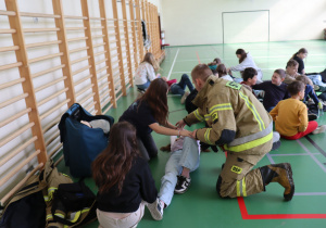 Uczniowie klasy 5a i 5b aGrupa dzieci oraz młodzieży uczestniczy w szkoleniu z pierwszej pomocy na sali gimnastycznej. Na pierwszym planie strażak w ubraniu ochronnym (z napisem „OSP”) wraz z dwiema nastolatkami udzielają pomocy osobie leżącej na podłodze, trzymając jej nogę. W tle inne dzieci obserwują lub ćwiczą podobne działania w parach. Wzdłuż lewej ściany widać drewniane drabinki gimnastyczne.z pomocą strażaka wykonują ćwiczenie ułożenie poszkodowanego w pozycji bocznej bezpiecznej.