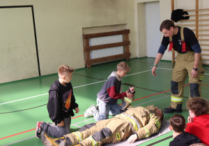 Na sali gimnastycznej dzieci uczą się pierwszej pomocy. Dwóch chłopców klęczy obok leżącego na plecach strażaka w mundurze ochronnym. Jeden z chłopców trzyma jego ramię, drugi obserwuje. Obok stoi inny strażak, nadzorujący ćwiczenie. W tle widoczna jest drabinka gimnastyczna, kurtka strażacka i grzejnik.