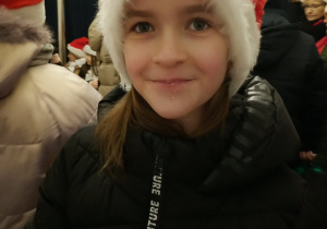Zbliżenie na uśmiechniętą dziewczynkę w czarnej kurtce i czerwonej czapce Mikołaja wśród tłumu osób zgromadzonych przy scenie jarmarku.