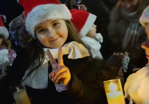 Uśmiechnięta uczennica SP 13 w czapce Mikołaja pokazuje znak pokoju i trzyma świecę podczas jarmarku.