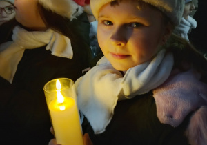 Dziewczynka z SP 13 w czapce Mikołaja trzyma zapaloną świecę podczas wieczornego fragmentu jarmarku.