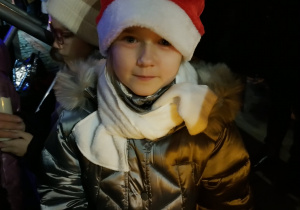 Dziewczynka w srebrnej kurtce, białej chuście i czerwonej czapce Mikołaja trzyma w dłoniach świecę podczas wieczornego Pabianickiego Jarmarku Świątecznego.