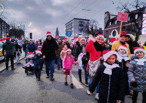 Grupa dzieci i dorosłych w zimowych kurtkach i czapkach Mikołaja idzie środkiem ulicy w świątecznej paradzie, część uczestników macha do obiektywu.