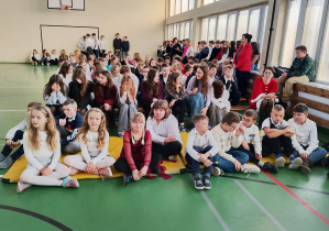 Uczniowie klas 4–8 siedzą w sali gimnastycznej podczas szkolnej uroczystości.