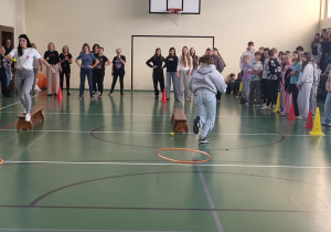 Uczennica i nauczycielka pokonują tor sprawnościowy w sali gimnastycznej, a zgromadzeni uczniowie i nauczyciele kibicują z boku.
