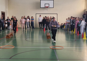 Uczennica pokonuje tor sprawnościowy w sali gimnastycznej, a uczniowie i nauczyciele kibicują.