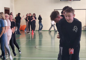 Uczniowie i nauczyciele wykonują wspólnie konkurencję drużynową w sali gimnastycznej.