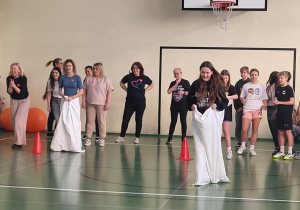 Uczniowie biorą udział w wyścigu w workach w sali gimnastycznej, a nauczyciele i rówieśnicy kibicują.