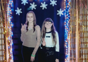 Dwie uczennice klasy siódmej pozują na tle karnawałowych dekoracji z balonami, śnieżynkami i lampkami. Obie stoją obok siebie na tle czarnego materiału, uśmiechnięte i pokazujące znak V rękami.